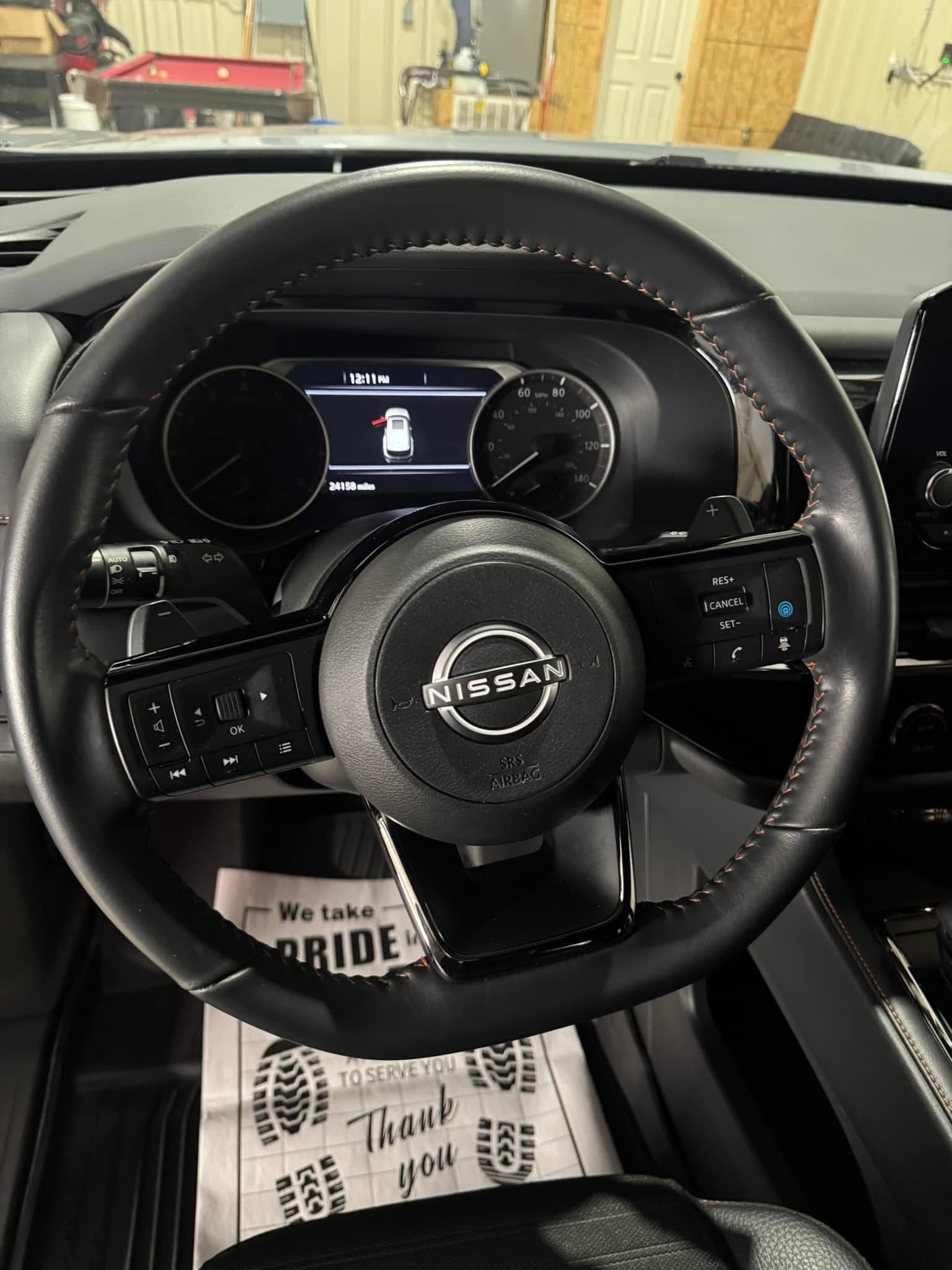 Black steering wheel in a Nissan vehicle, with dashboard visible.