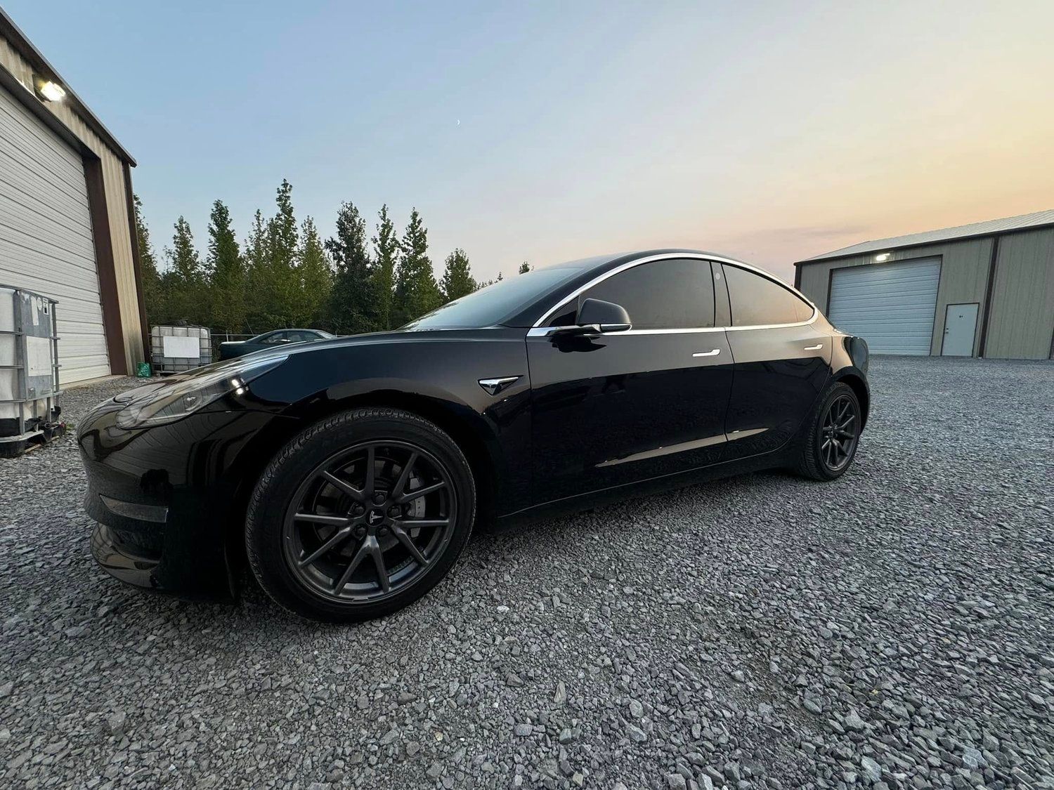 Black Tesla Model 3 parked on gravel, beside a building.