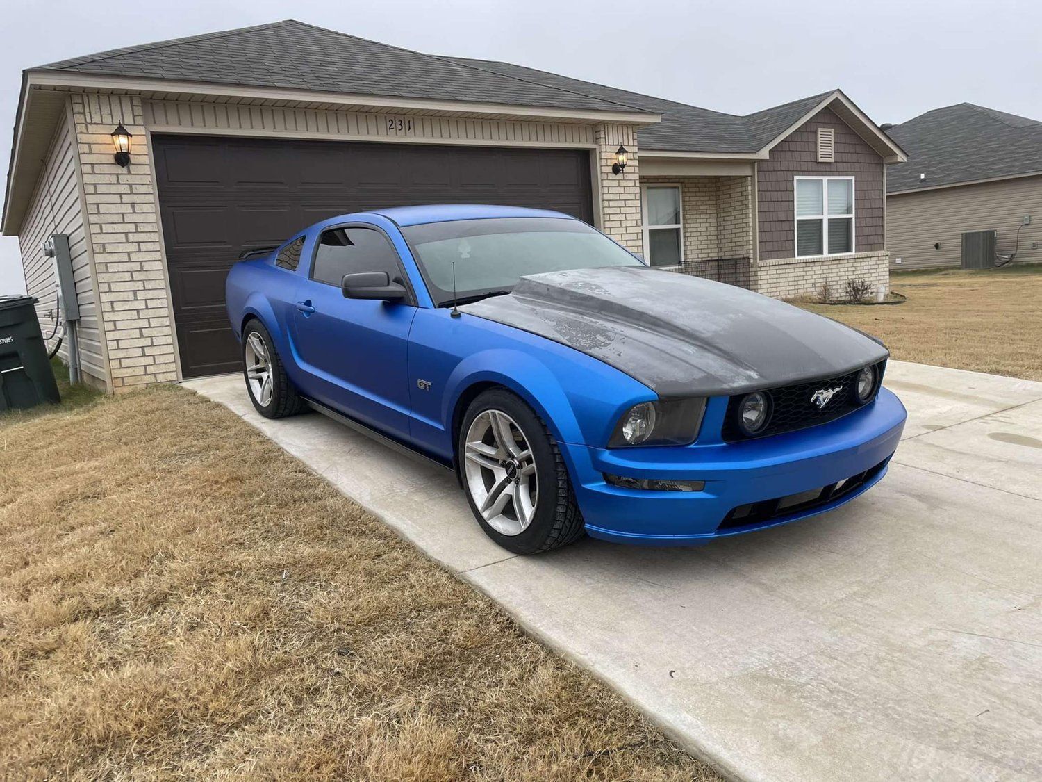 Blue Ford Mustang parked in a driveway in front of a house. Hood is black, setting is overcast.