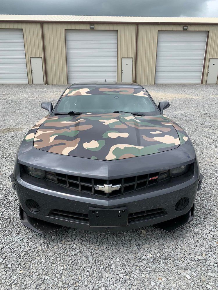 Camouflaged Chevrolet Camaro parked in front of garage doors, gray and black paint scheme.