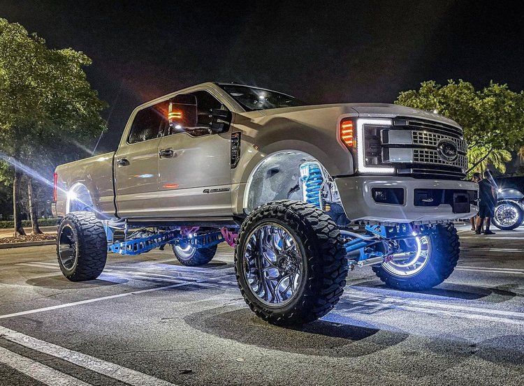 Lifted white Ford truck with large chrome wheels, LED lights, parked at night.