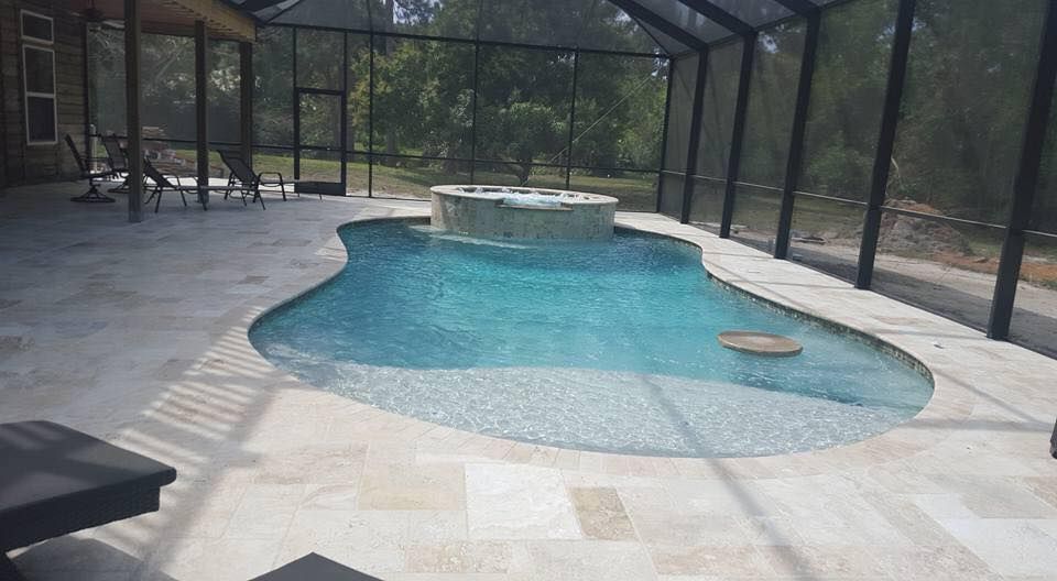 Swimming pool and spa in a screened enclosure, light blue water, light tile decking, surrounded by trees.