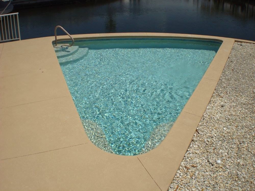 Triangular swimming pool with curved side, light blue water, beige concrete deck, and gravel border.