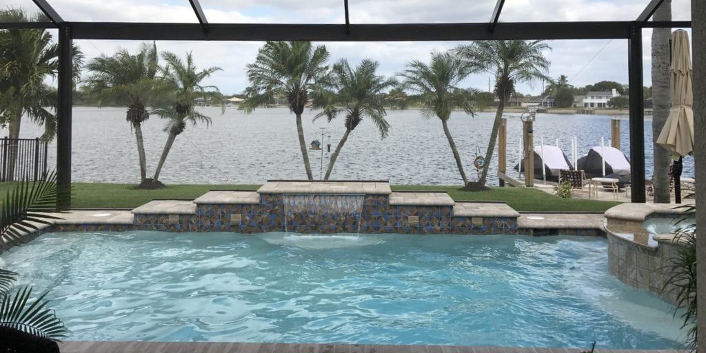 Pool overlooking a lake with palm trees. Grey patio, blue water.