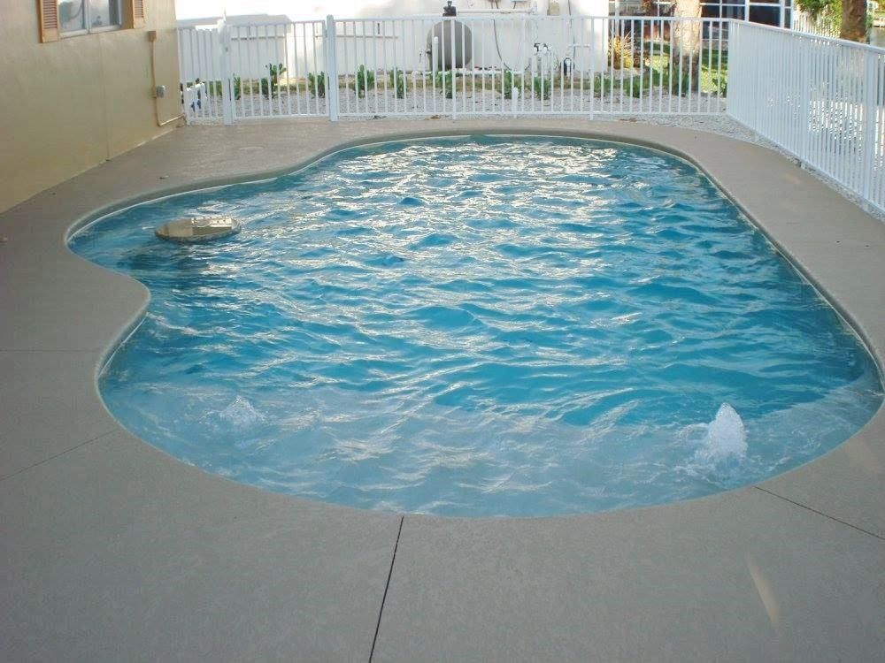 Small, kidney-shaped swimming pool with blue water, surrounded by a concrete deck and white fence.