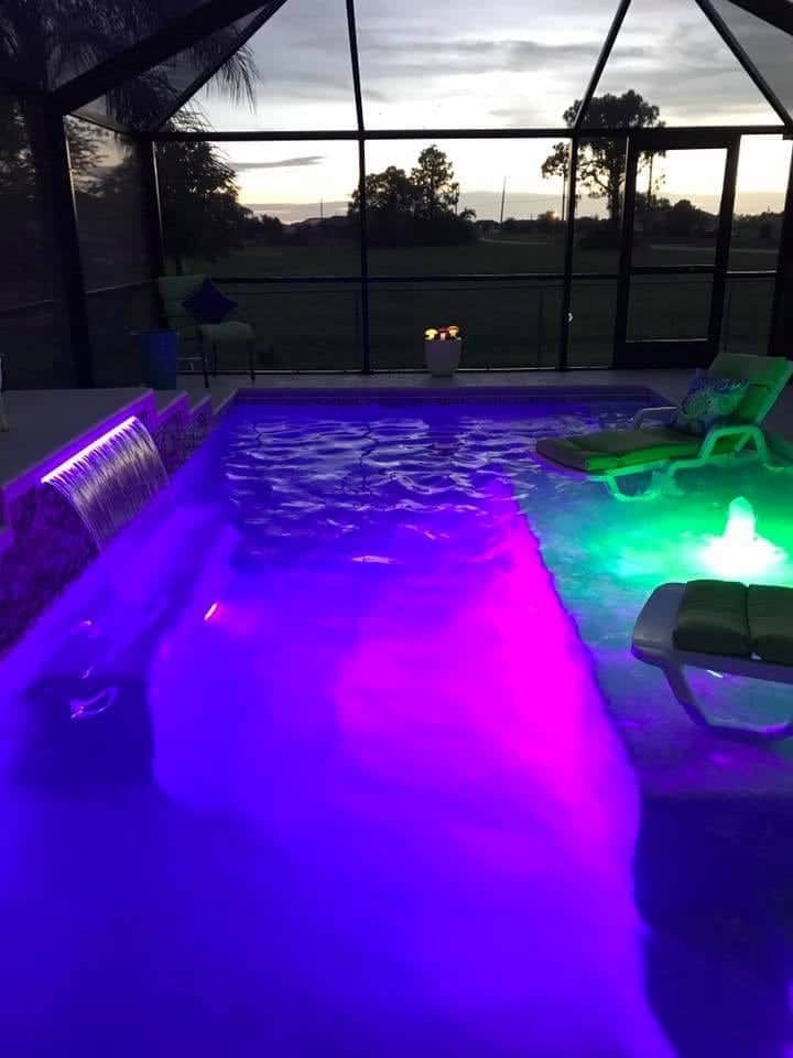Pool illuminated with purple and green lights, surrounded by a screened-in patio at dusk.
