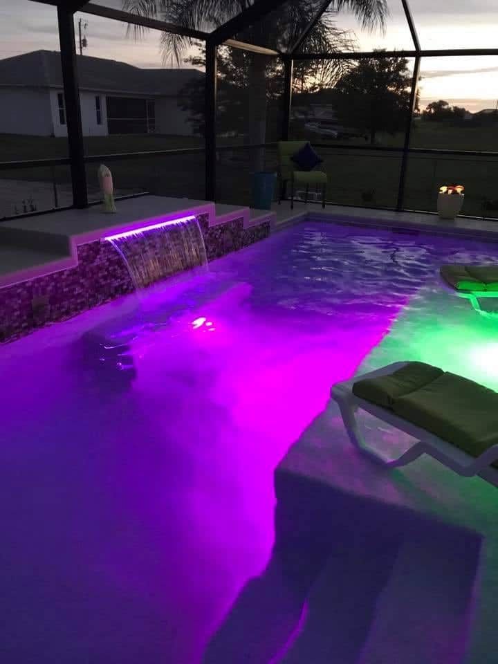Pool lit with purple and green lights, waterfall feature, patio furniture, screened enclosure, evening setting.