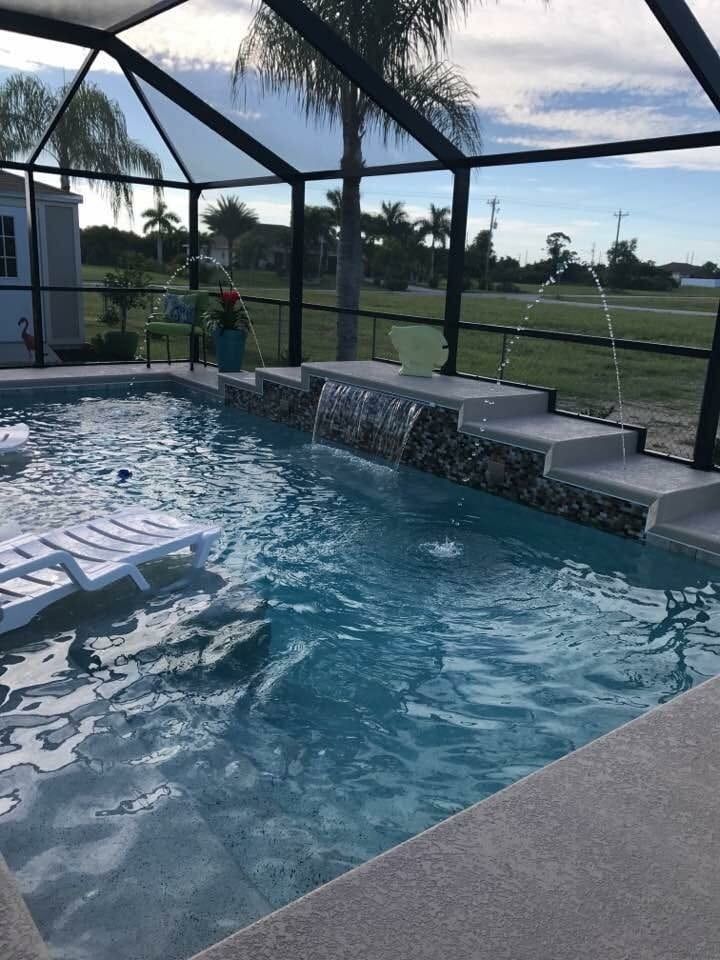 A pool with water features and a lounging chair, enclosed by a screen, with a view of a green field.
