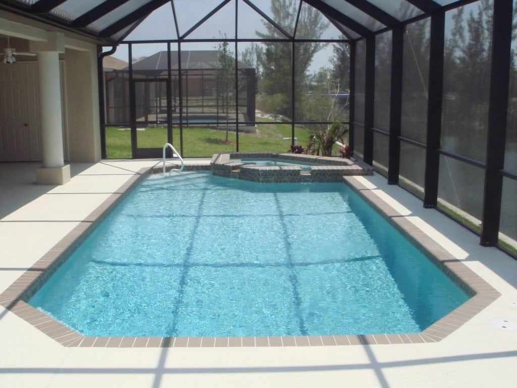 A screened-in pool with turquoise water, a small hot tub, and a view of a backyard.