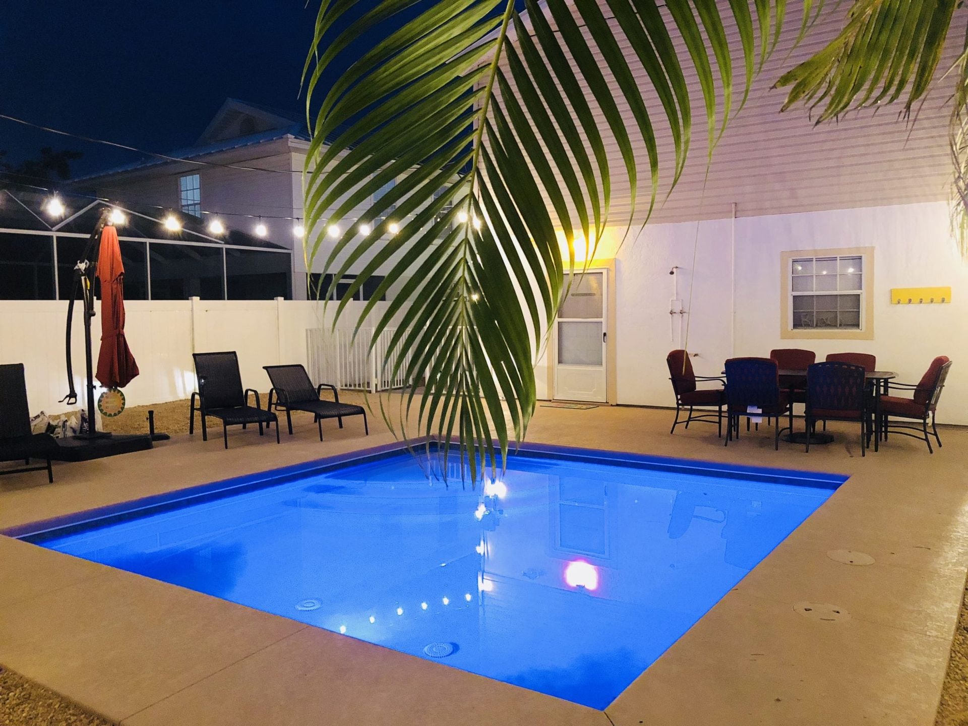 Nighttime pool area with illuminated blue water, lounge chairs, a dining table, and palm fronds.