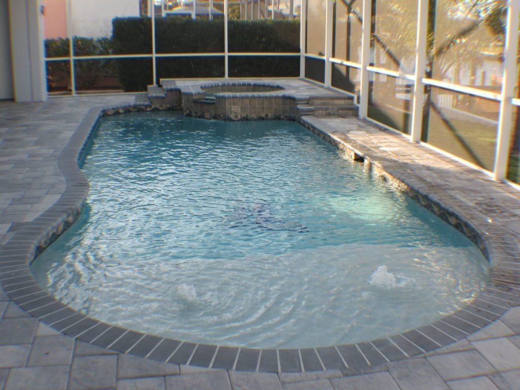 Pool with a spa, surrounded by brick pavers, screened enclosure. Water features and landscaping visible.