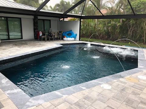 Pool with fountains, patio, and screened enclosure; blue water, grey pavers, and lush greenery in background.