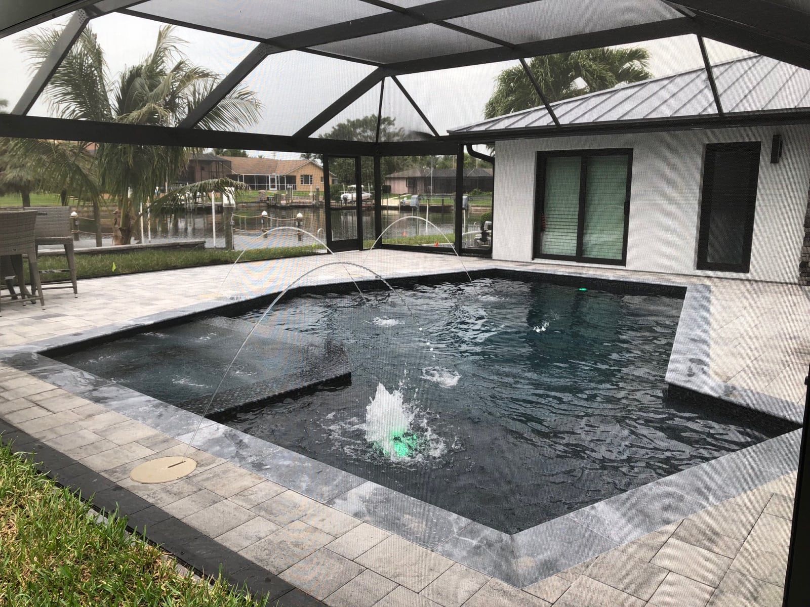 Pool enclosed in a screen. Gray pavers surround the pool with fountains. White home in background.