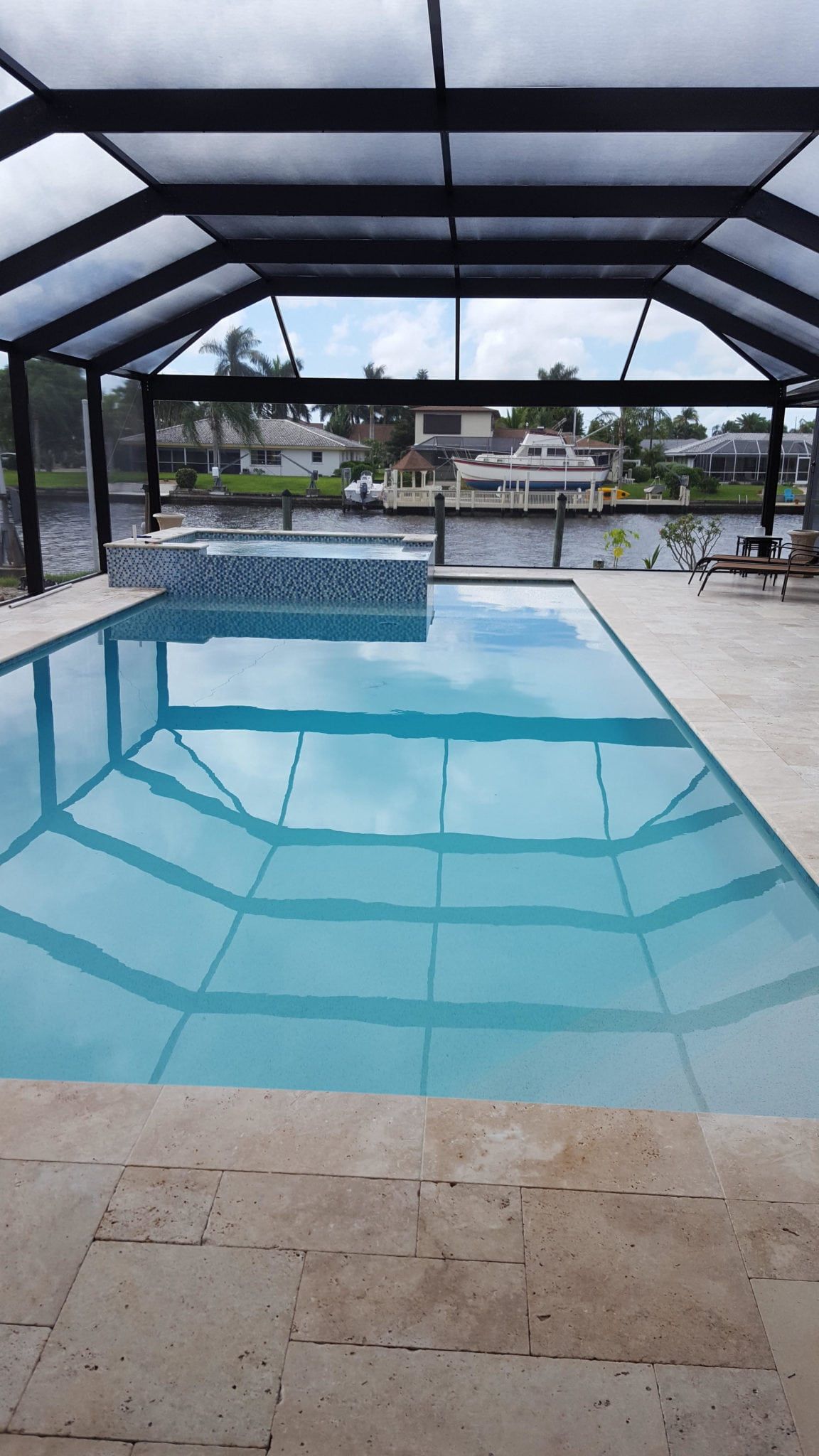 A rectangular pool with blue water, enclosed in a black screened structure, overlooks a canal with houses.