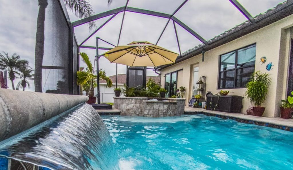 A pool with a waterfall feature, umbrella, and tropical plants under a screened enclosure.
