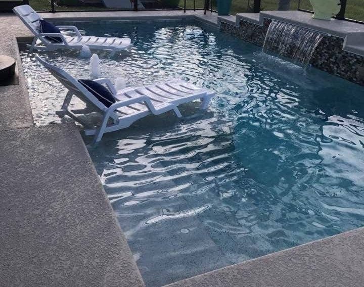 Pool with two white lounge chairs in shallow water, waterfall feature, and gray concrete surround.