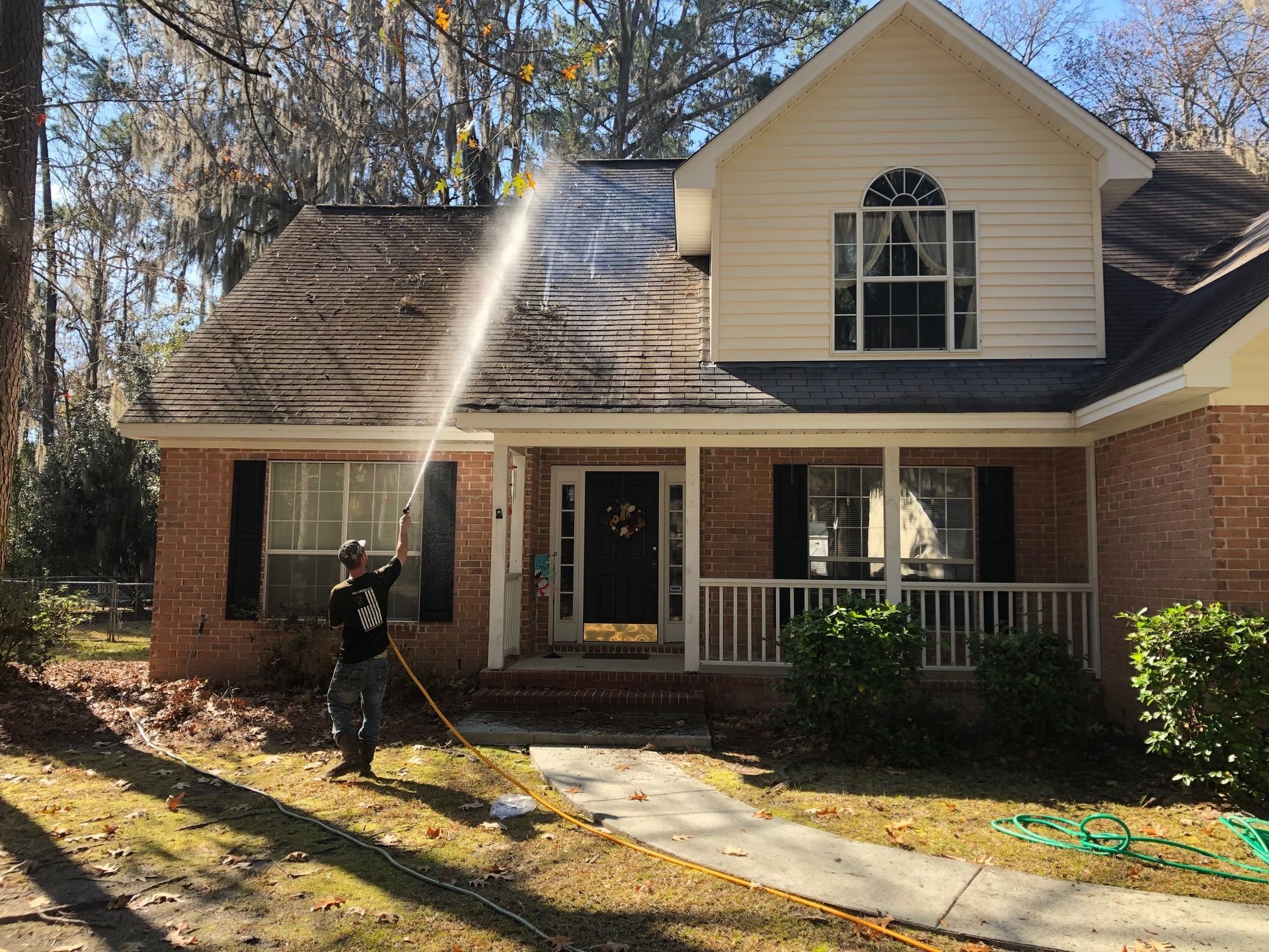 Man pressure washing a two-story brick and white house with a dark roof on a sunny day.