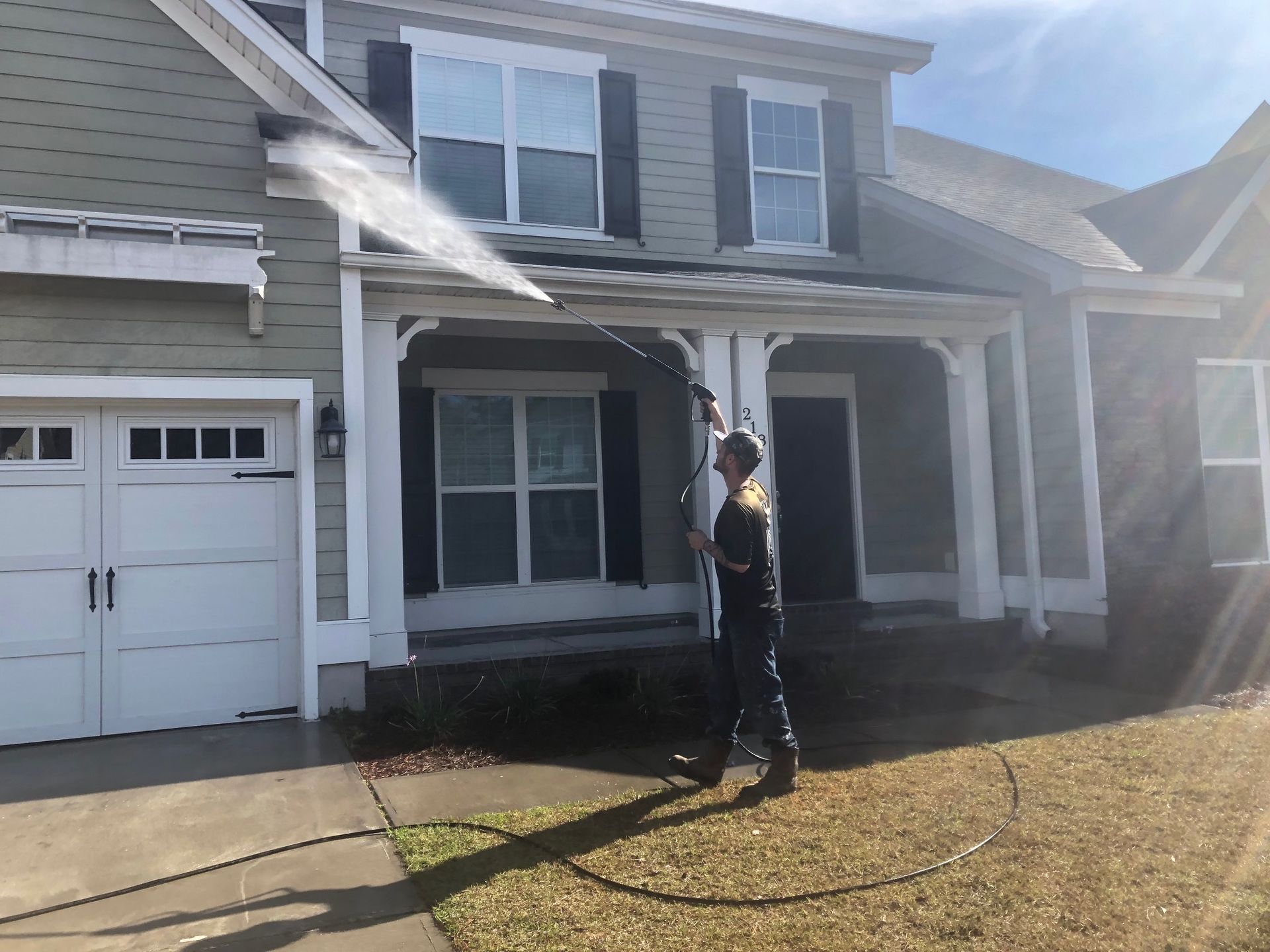 Man pressure washing a two-story house with a gray exterior, spraying the front porch on a sunny day.