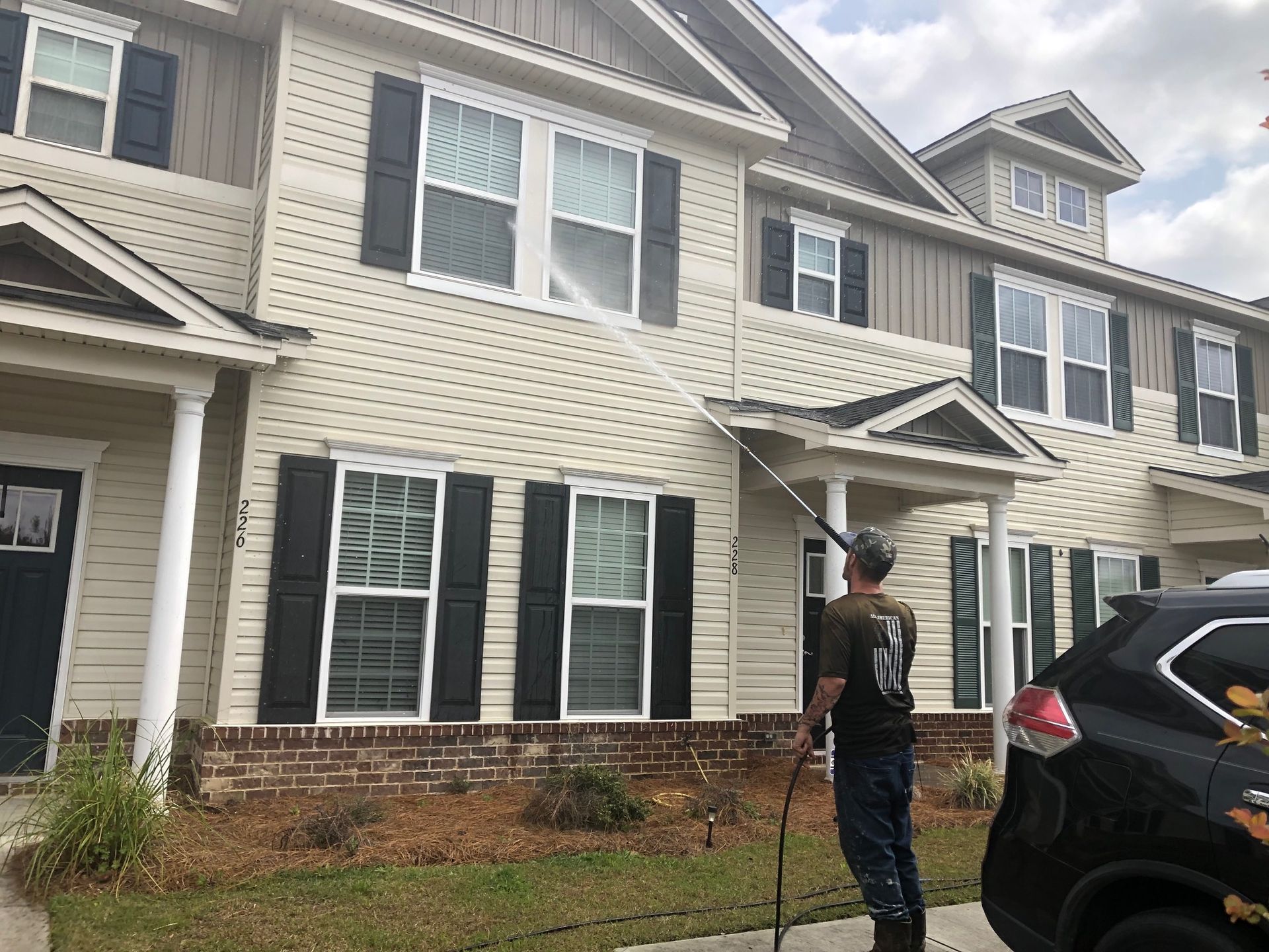 Man power washing the siding of a light tan townhouse. Black shutters, blue sky, and a black car in the foreground.