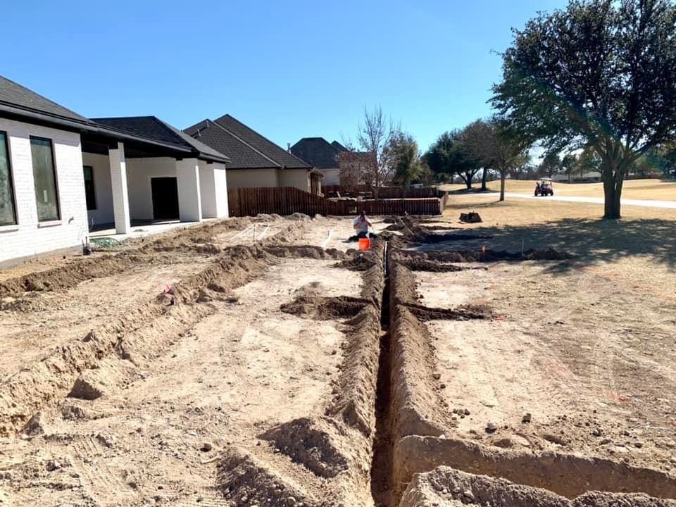 Trenches dug in dirt next to a house and golf course. Construction worker in the distance. Sunny day.