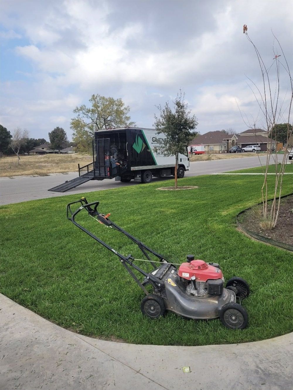 Lawnmower on green grass; landscaping truck in the background. Cloudy sky, suburban setting.