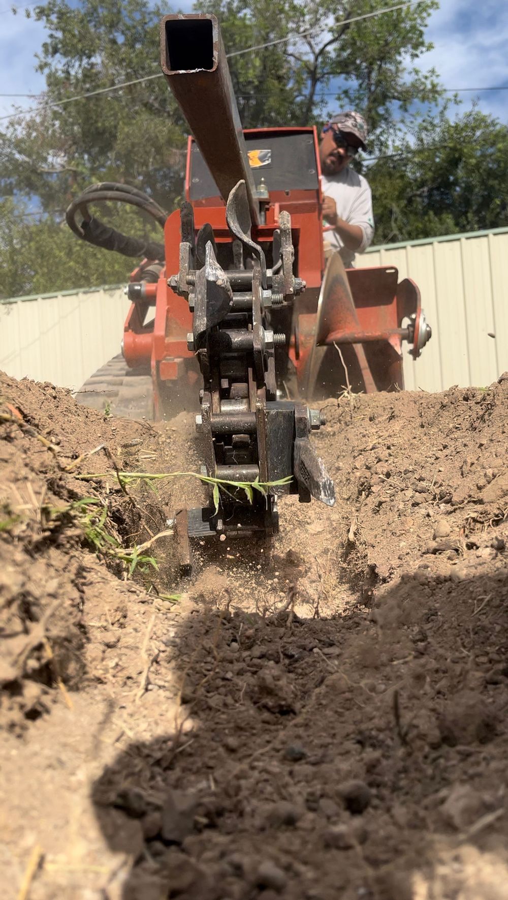 A man operates a ditch witch, digging a trench in brown soil near a building.