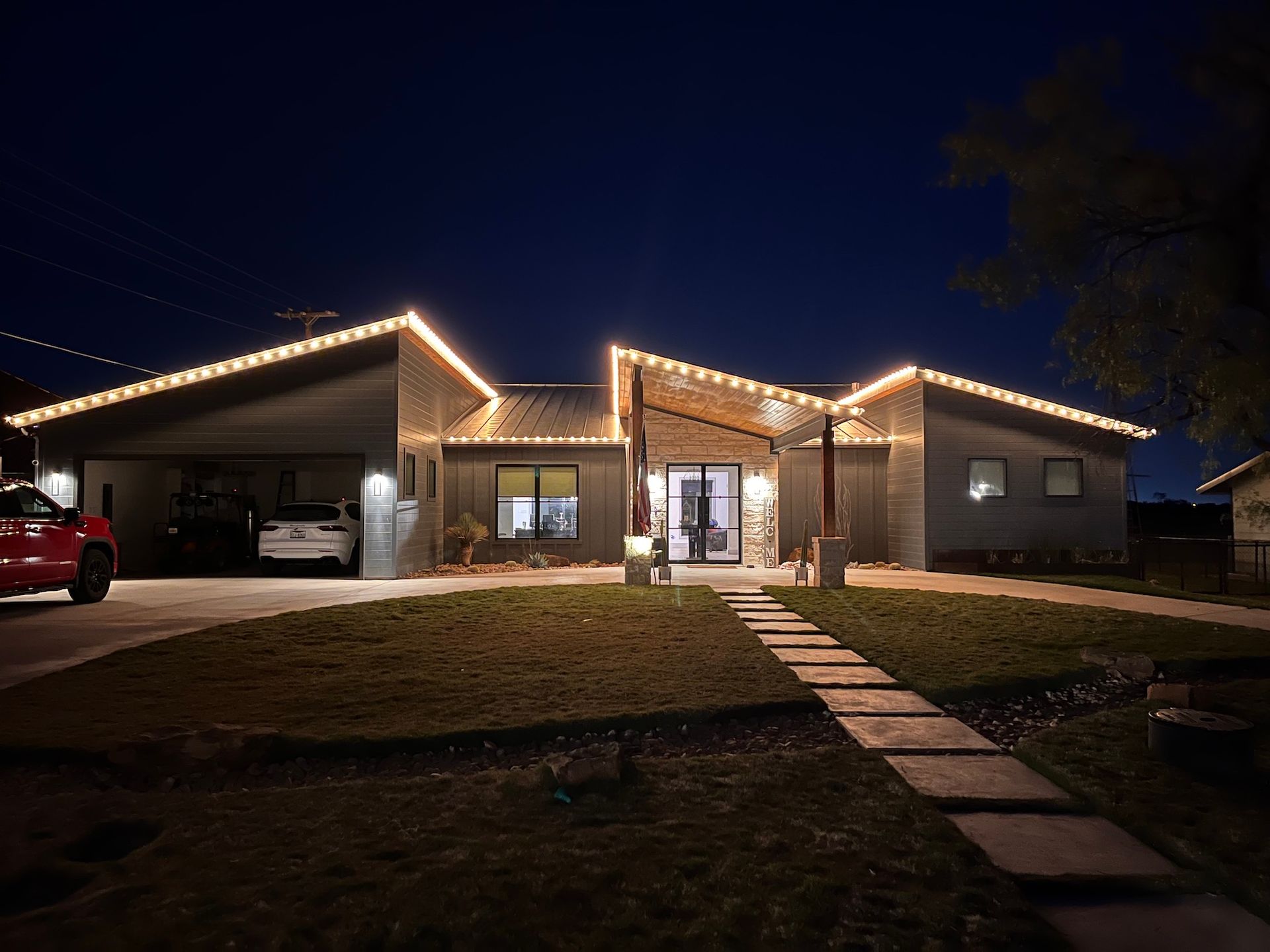 A house with holiday lights at night. A red car sits in the driveway, and a walkway leads to the front door.
