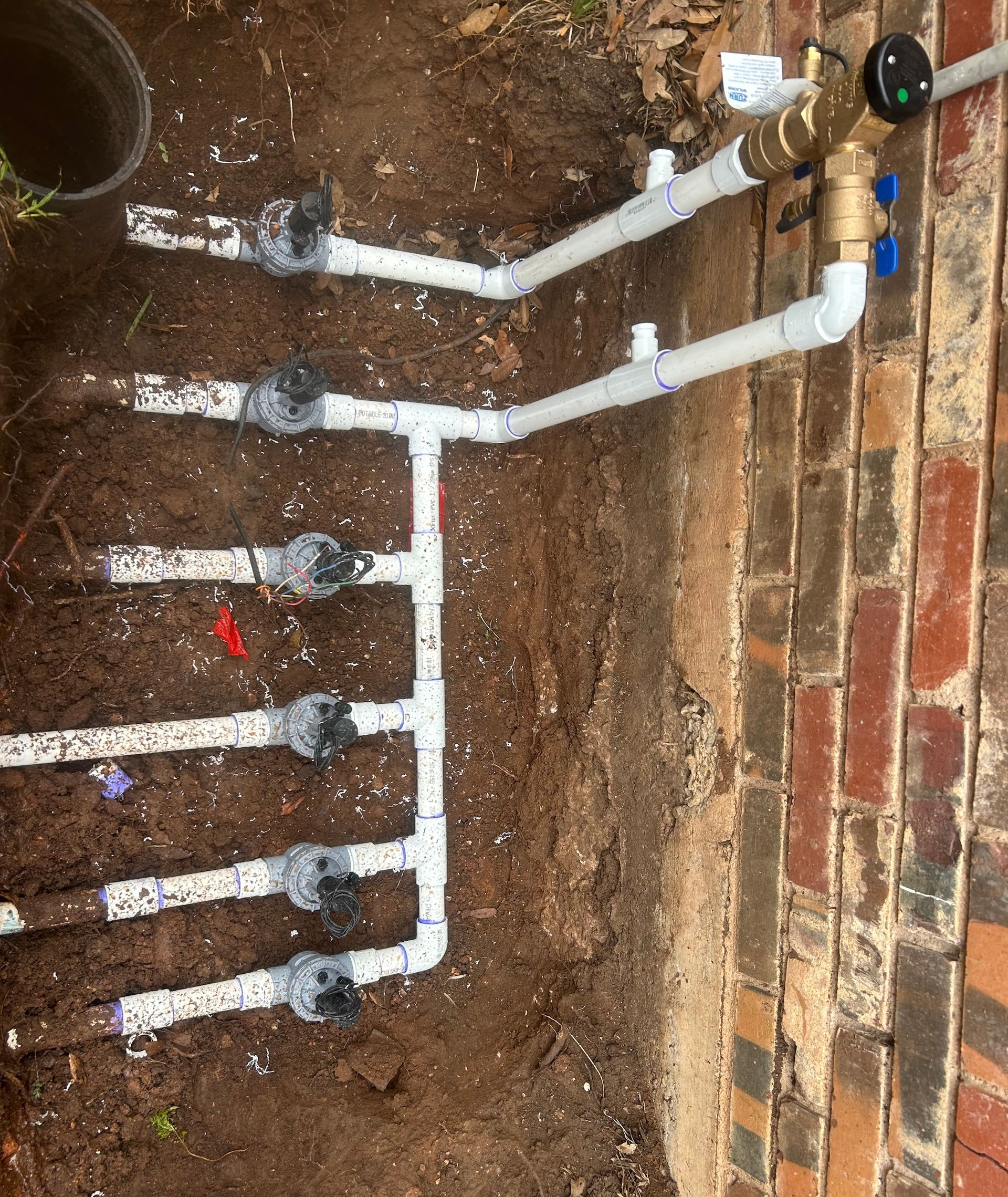 White PVC pipes and sprinkler valves on the ground near a brick wall, part of an irrigation system installation.