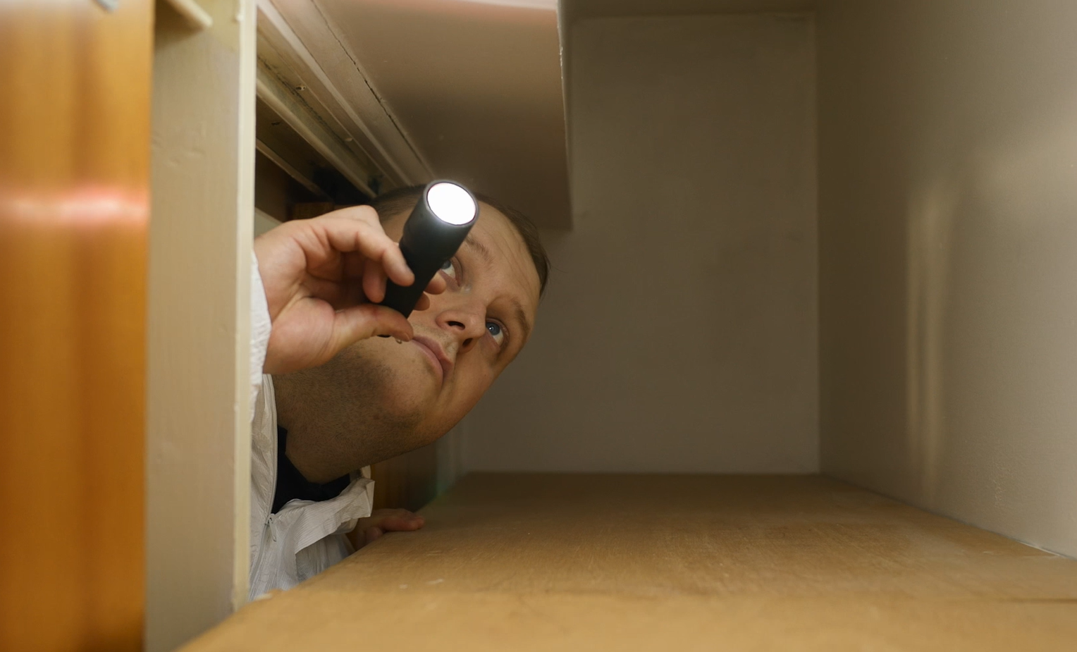 A man is peeking out of a closet with a flashlight.