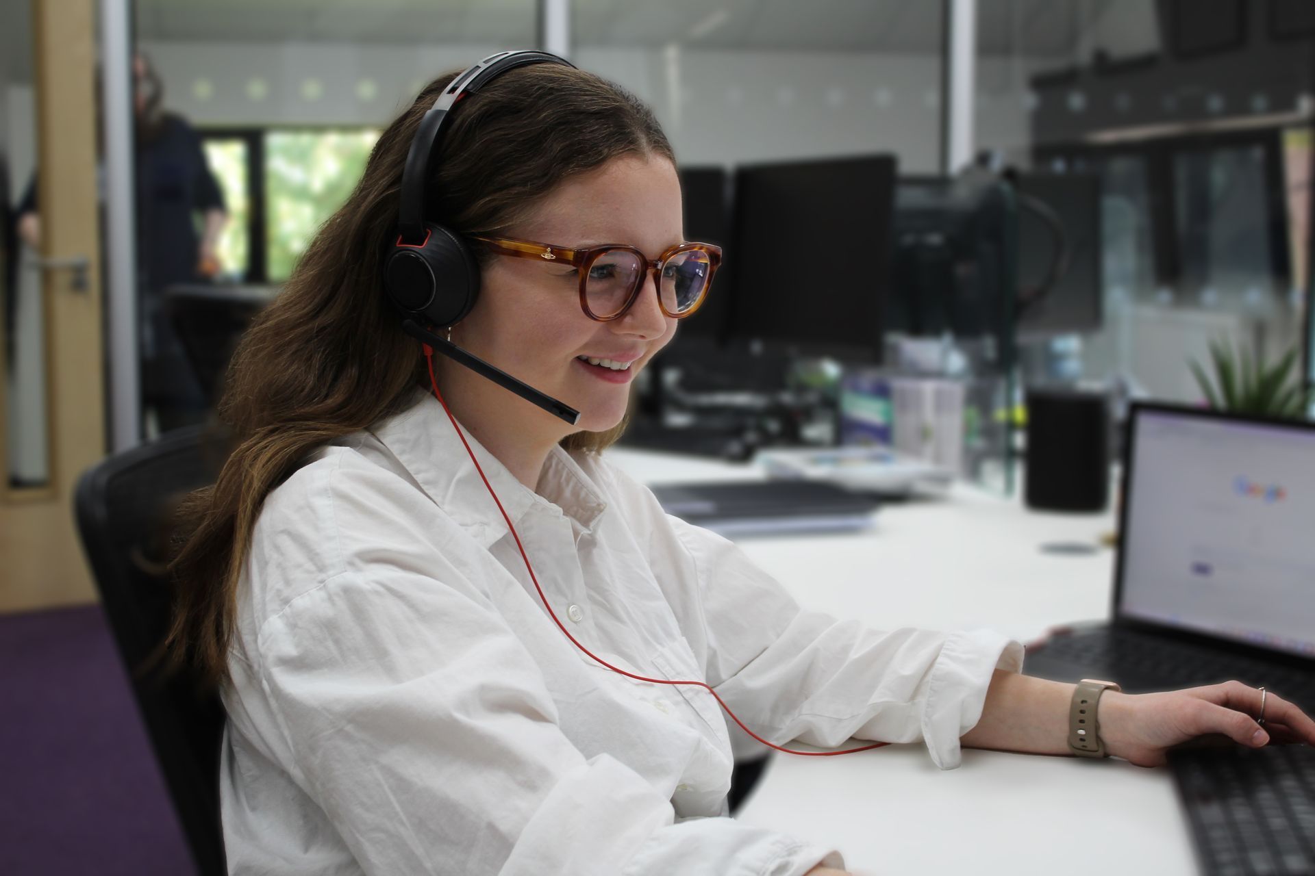 A woman wearing headphones and glasses is sitting at a desk in front of a laptop computer.