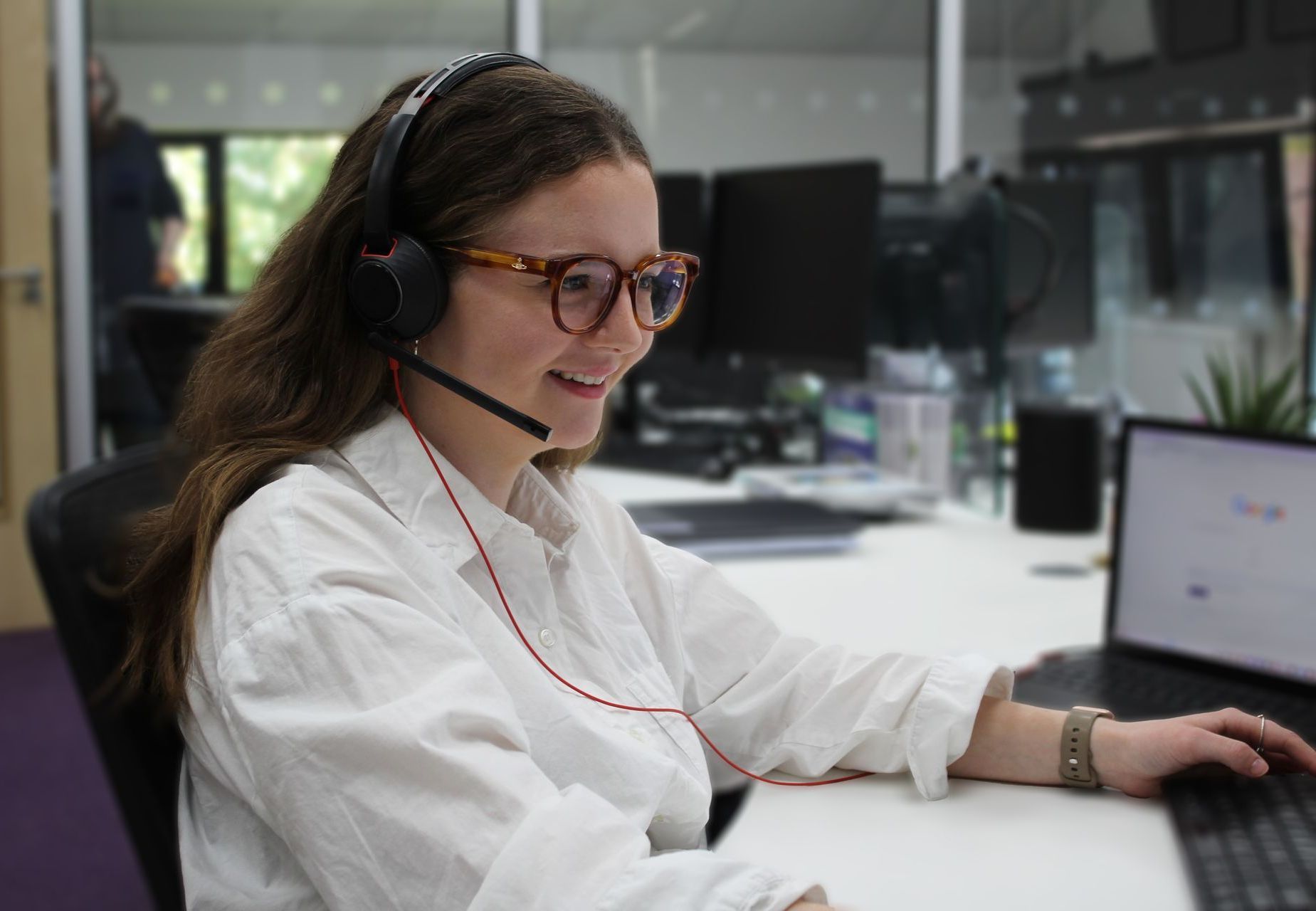 A woman wearing headphones and glasses is sitting at a desk in front of a laptop computer.
