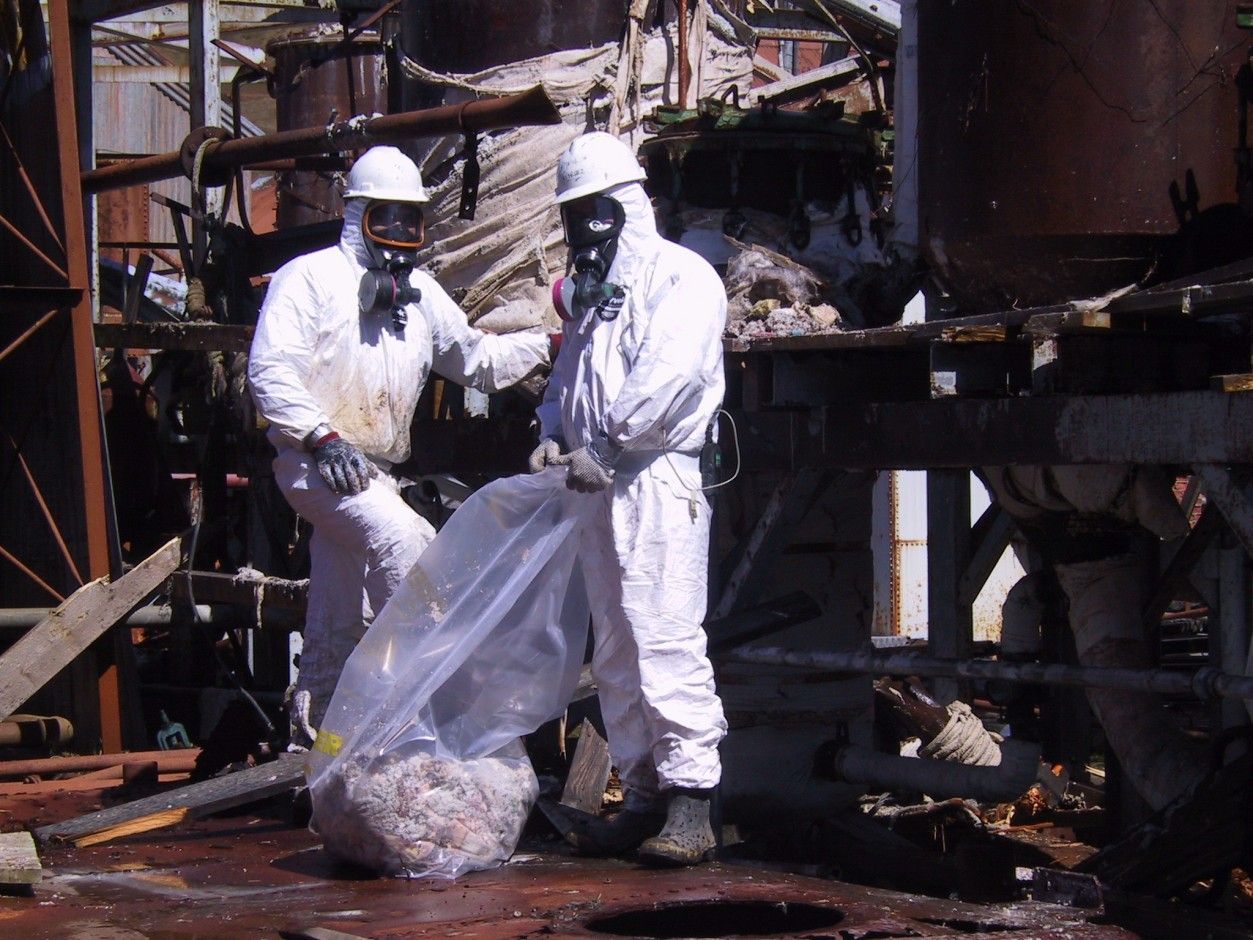 Two men wearing white hazmat suits, masks and hard hats are standing next to each other