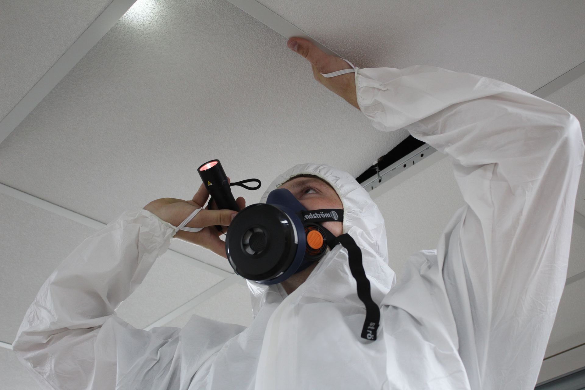 A man wearing a gas mask is looking up at the ceiling while holding a flashlight.