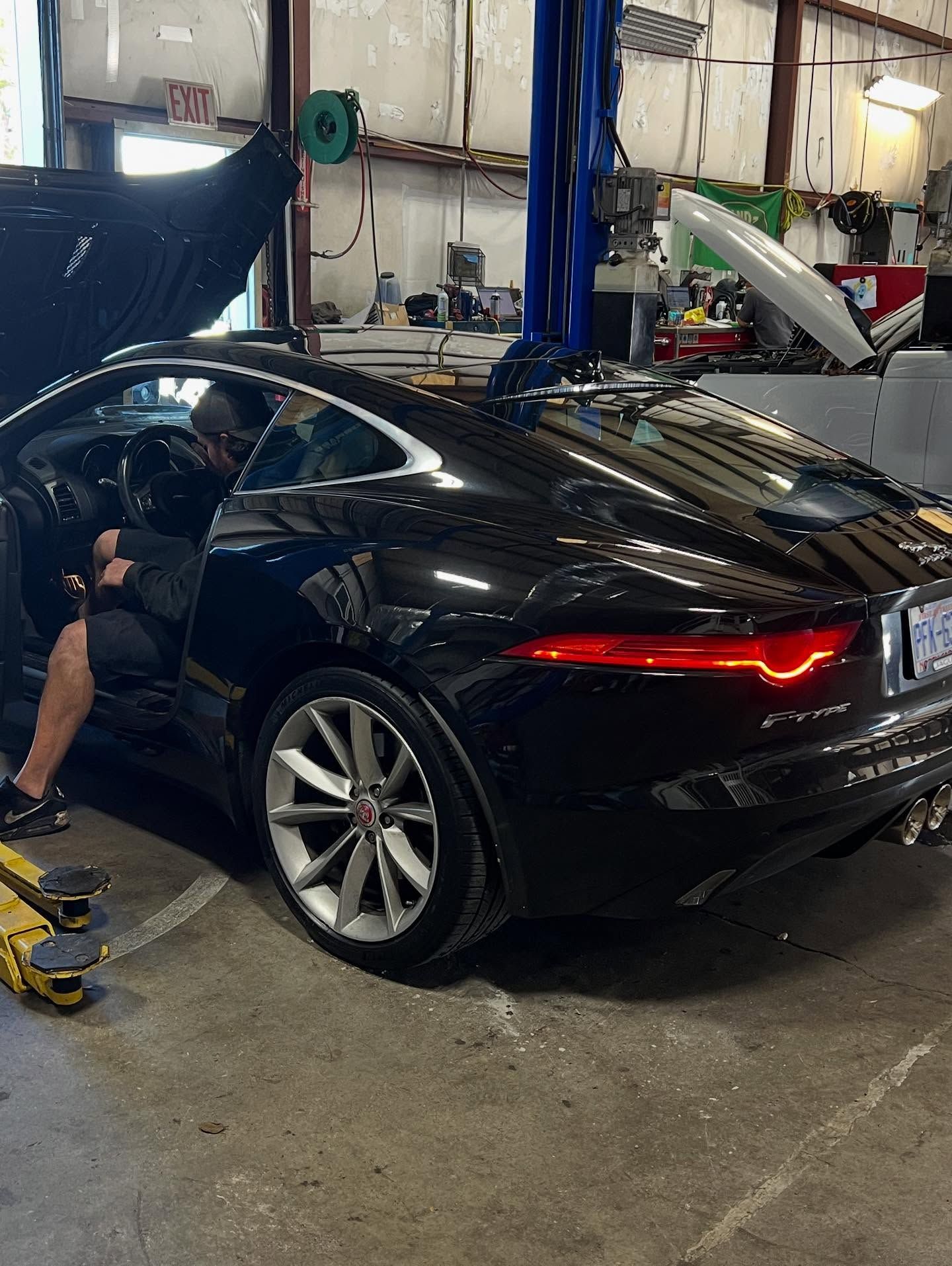 Black Jaguar F-Type in a repair shop with a person inside. Car is on a lift.