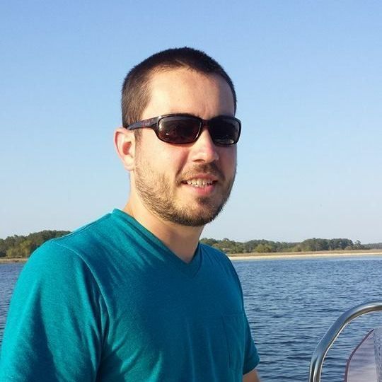 A man wearing sunglasses and a blue shirt is standing in front of a body of water