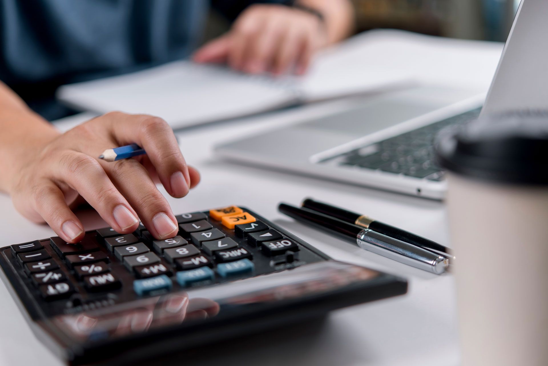 Person's hand using a calculator; laptop, notebook, pens, and coffee cup on a white desk.