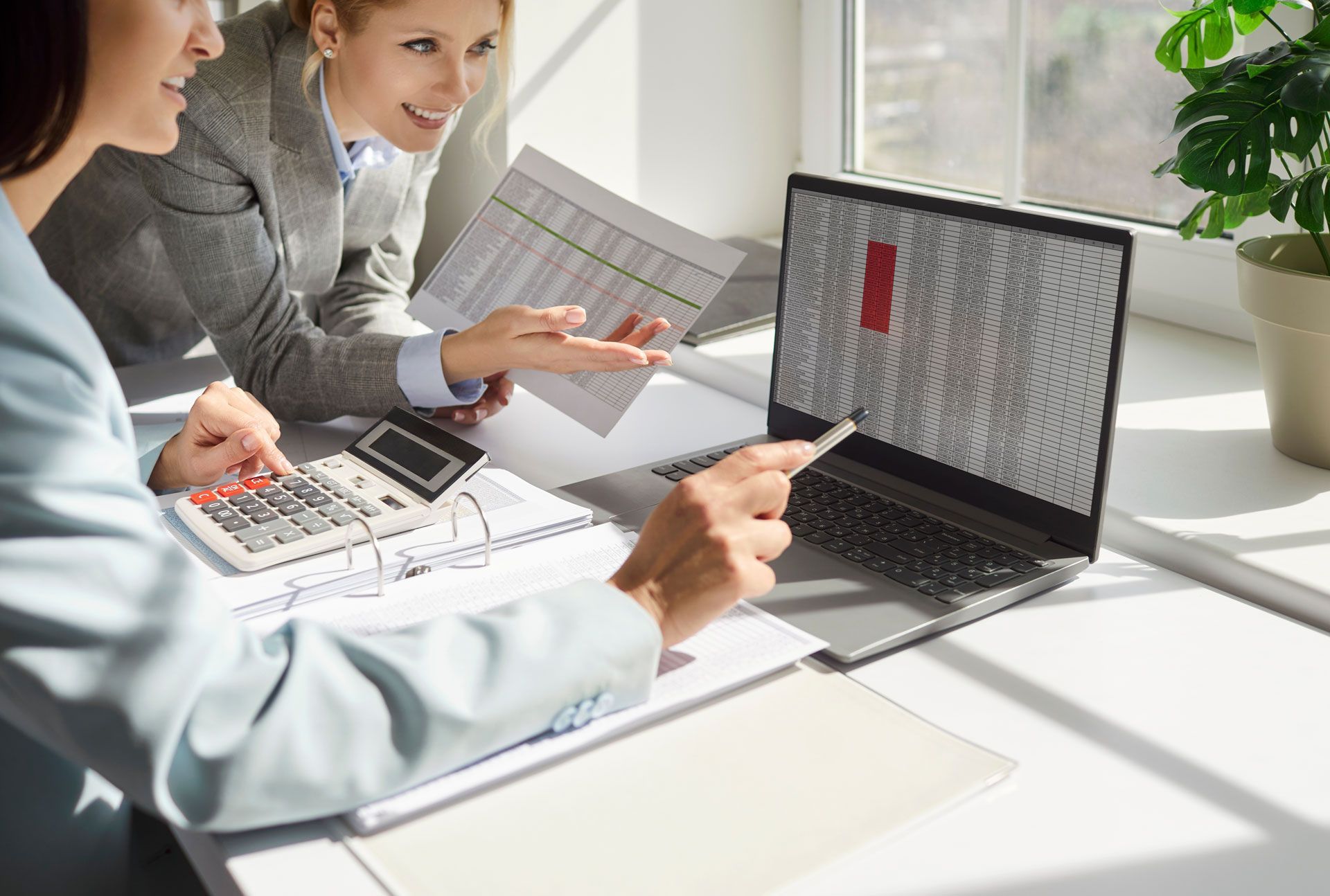 Two women reviewing financial data on a laptop and paper documents, using a calculator.
