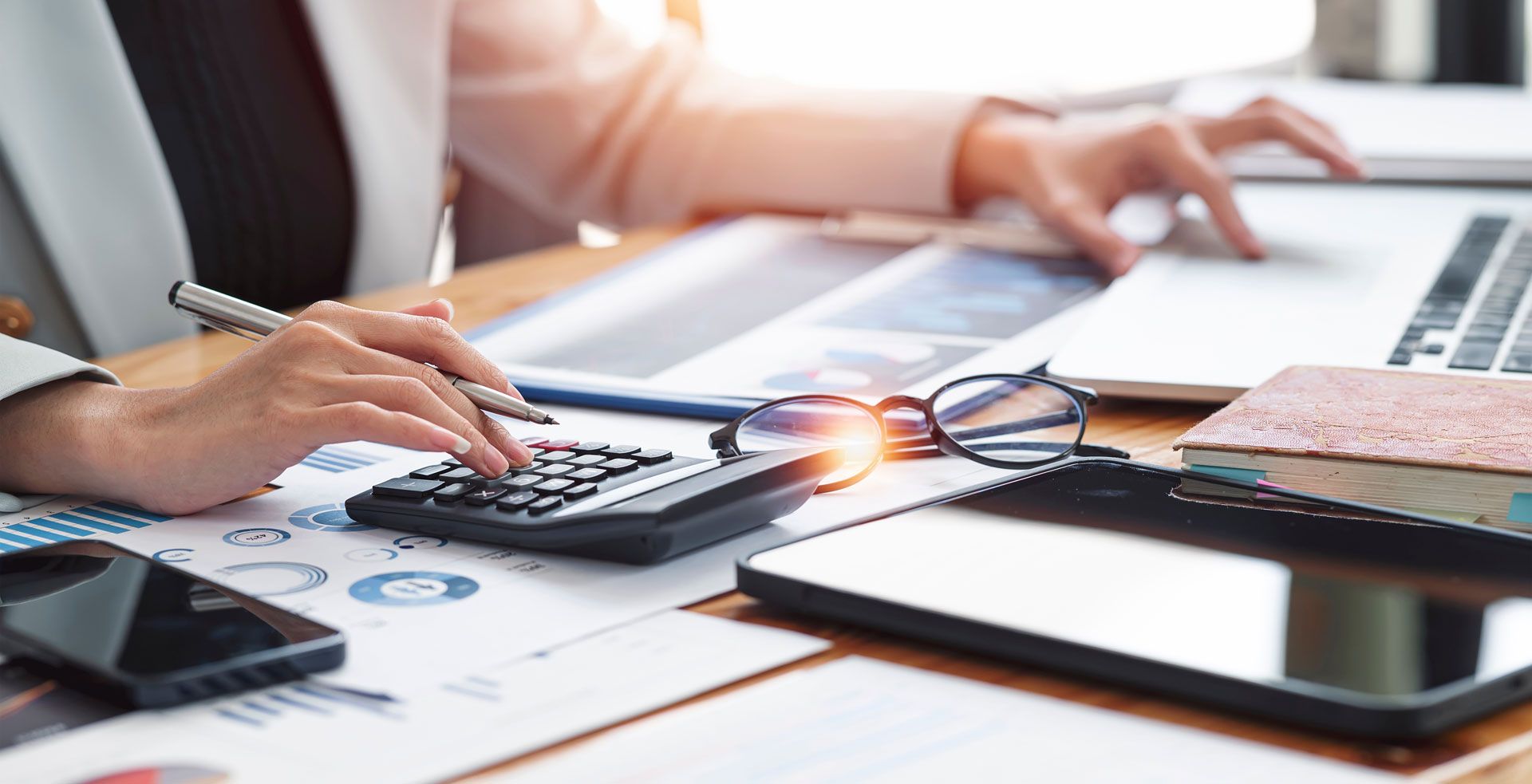Person using a calculator and laptop, working on financial documents at a desk with a smartphone, glasses, and a tablet.