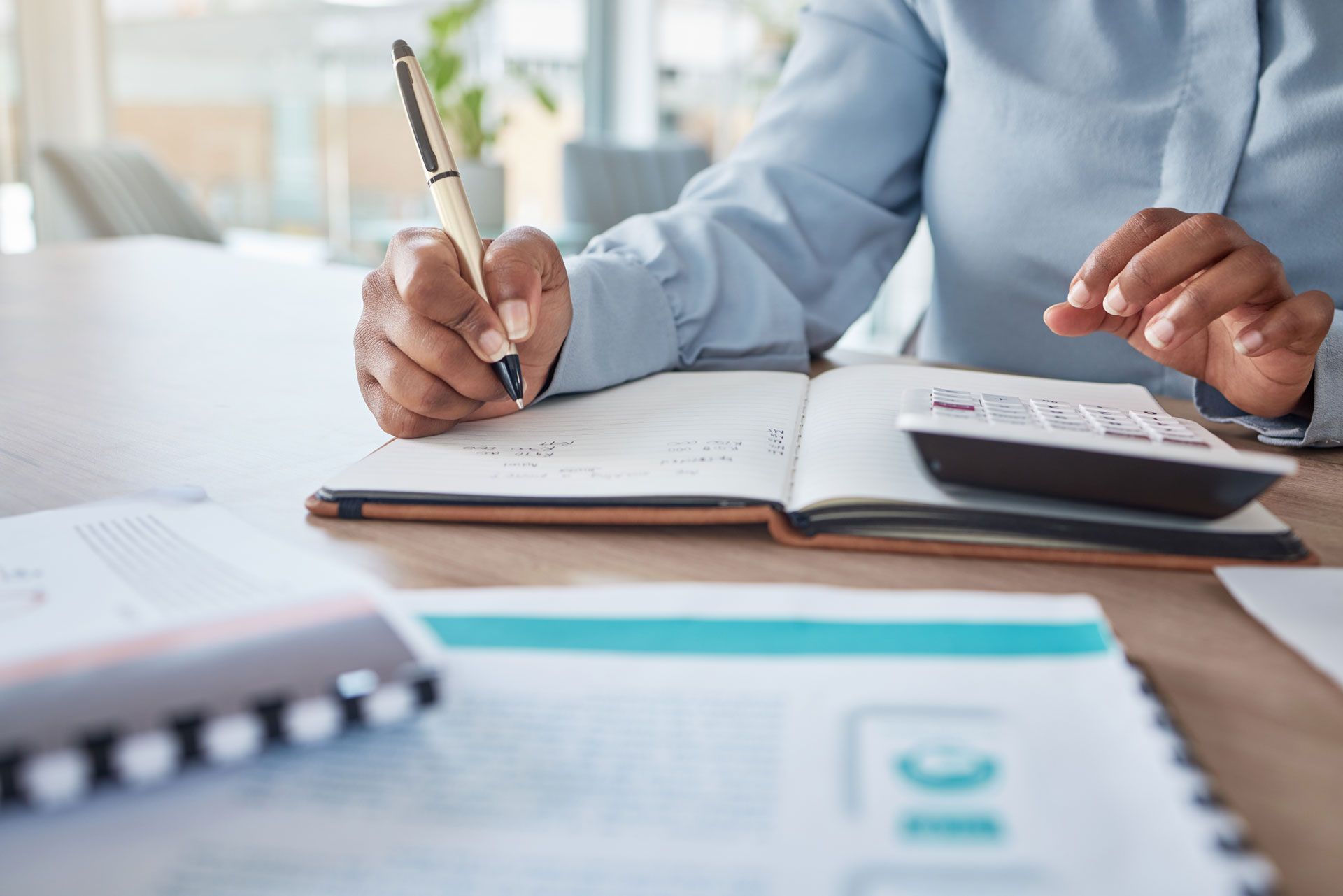 Person writing in notebook, using calculator at a desk, surrounded by documents.