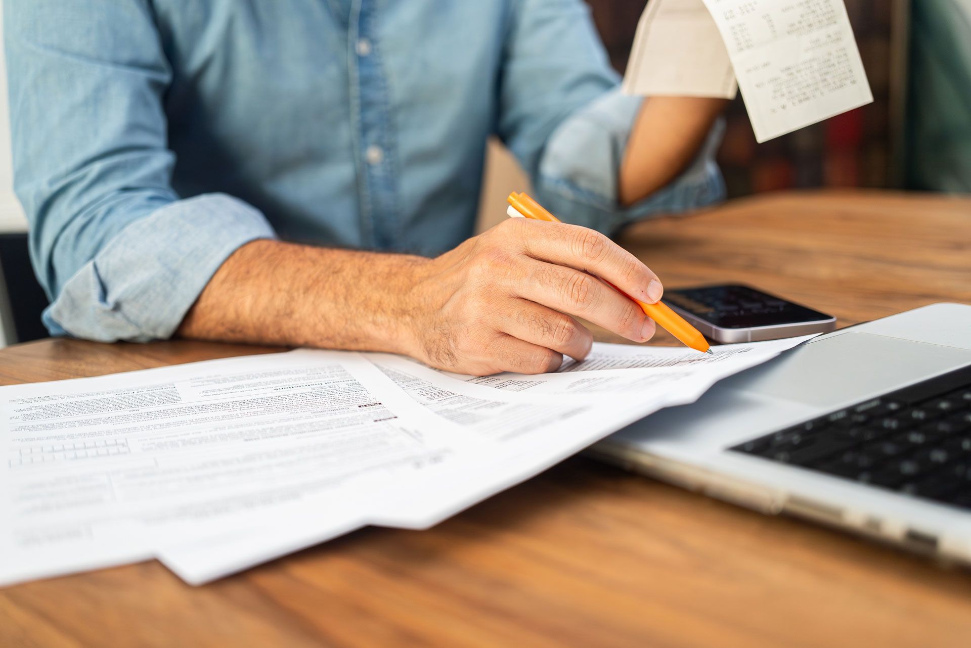 Person in blue shirt reviews receipts, papers, and laptop on wooden desk, using an orange pen.