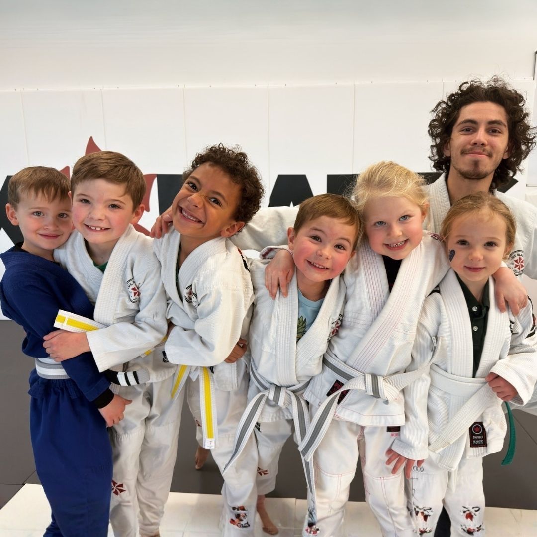 Five people in jiu-jitsu uniforms on a blue mat, including a man with a red belt, posing in a gym.