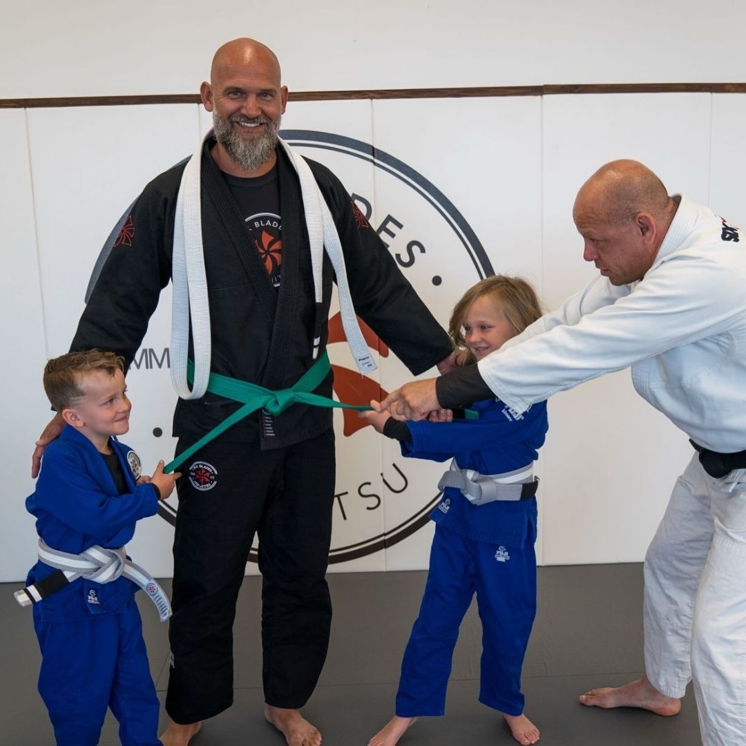 People practicing Brazilian Jiu-Jitsu. One person on the ground, another on top; two others grappling nearby. Black gis, indoor setting.
