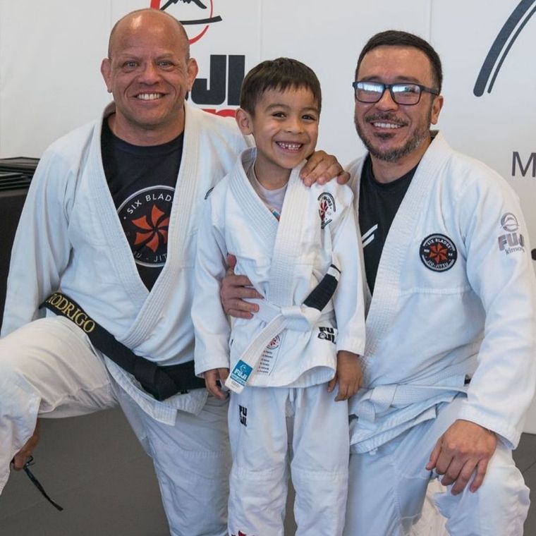 Family in Jiu-Jitsu gis poses. Indoors, blue mats, a smiling woman in the middle, around a seated man with a beard.