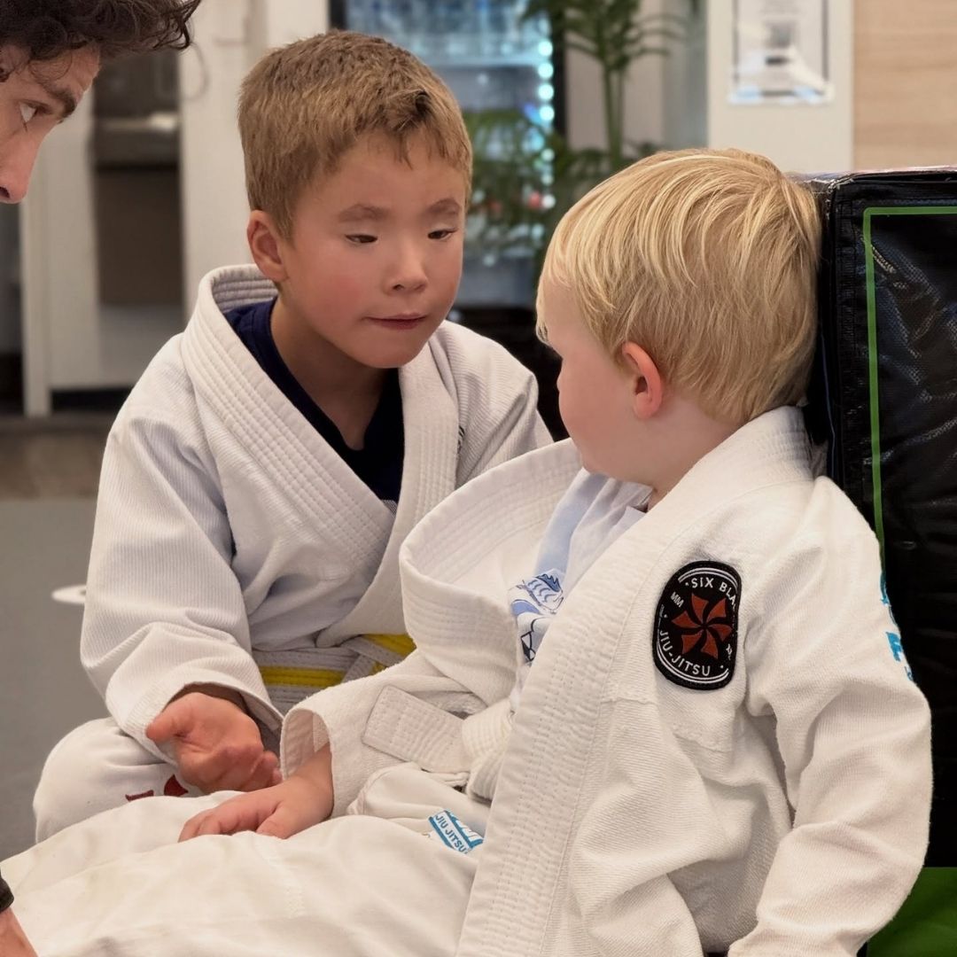 Children in colorful gis play a game on a mat. Two instructors pull a student in blue.