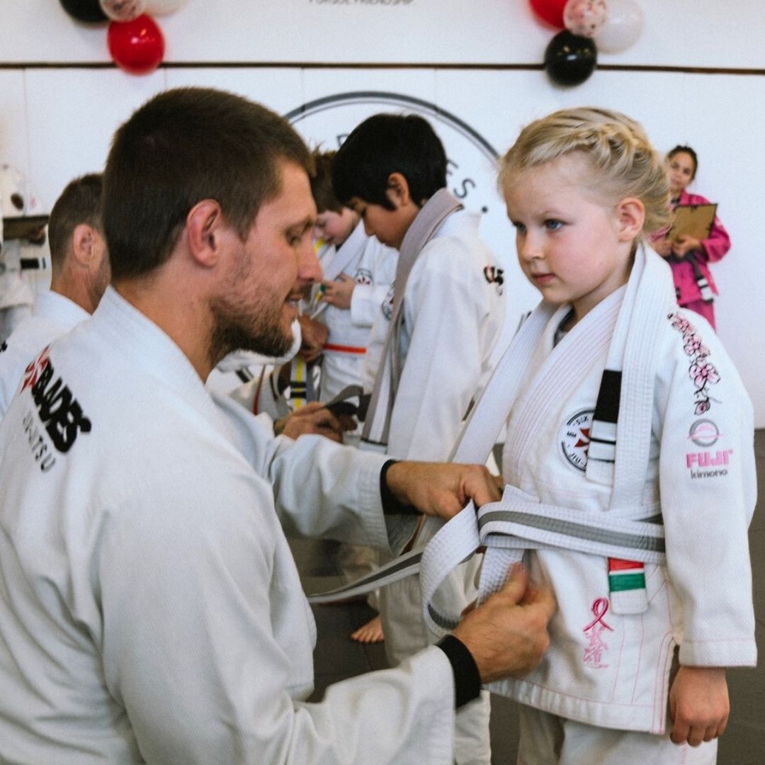Two children practicing Brazilian Jiu-Jitsu on a mat. One child in white gi has the other in a leg lock. A girl watches.