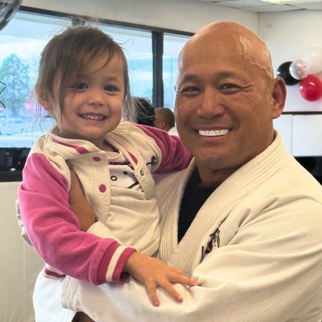 Two children in black and white martial arts uniforms practice a chokehold on a mat. One child smiles.