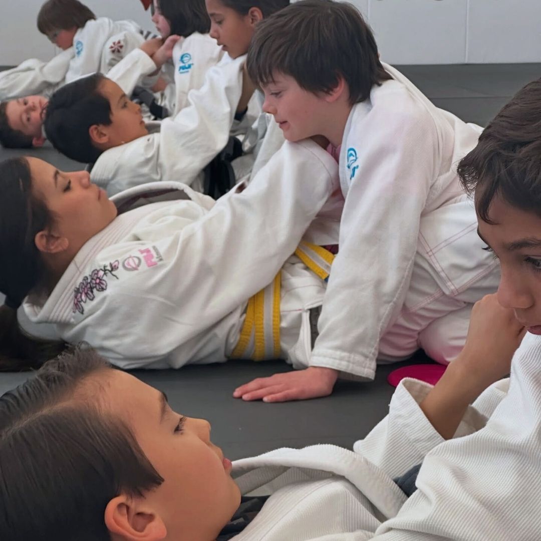 Two young children in blue and black martial arts uniforms grappling on a gym floor, one smiling.