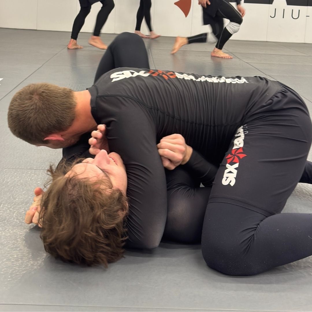 Five people in jiu-jitsu gear pose together in a gym.  Smiling,  some have arms around each other. Machado logo visible.