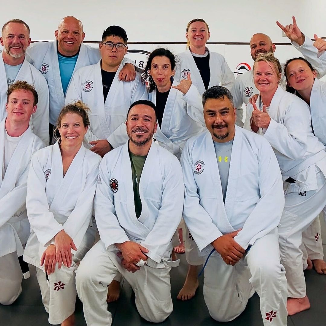 Two men in grappling position on a mat. One embraces the other from behind, both smiling. Gym setting.