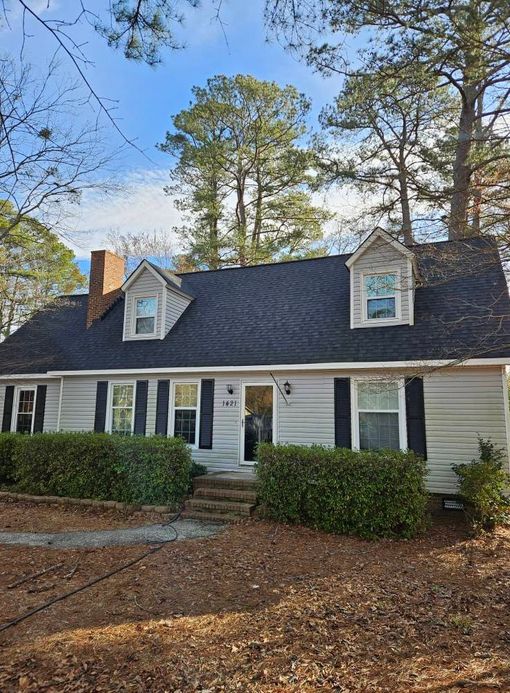 A gray house with black shutters, a brick chimney, and two dormers on a blue sky day.